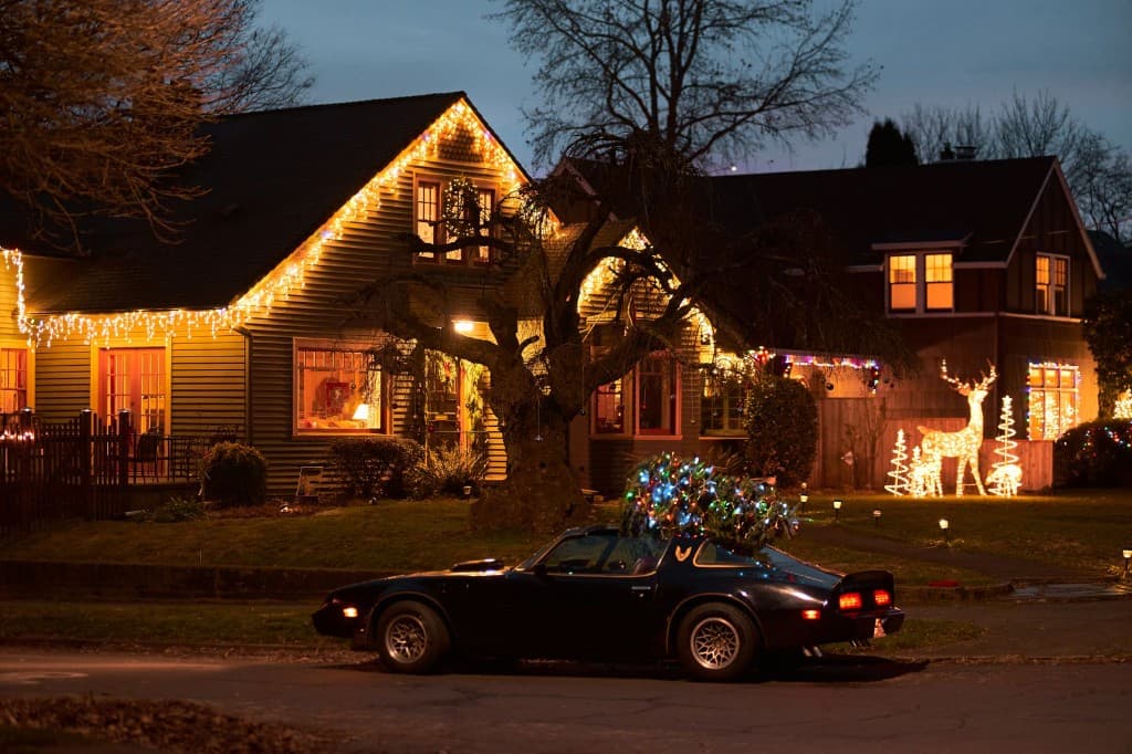 Yellow bungalow with warm white roofline lights and a Christmas tree on a parked car